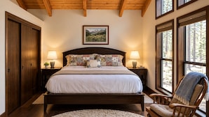 Quarto com cama grande, cabeceira de madeira, duas mesas de cabeceira com luminárias, uma cadeira de madeira e uma janela ampla com vista para árvores.