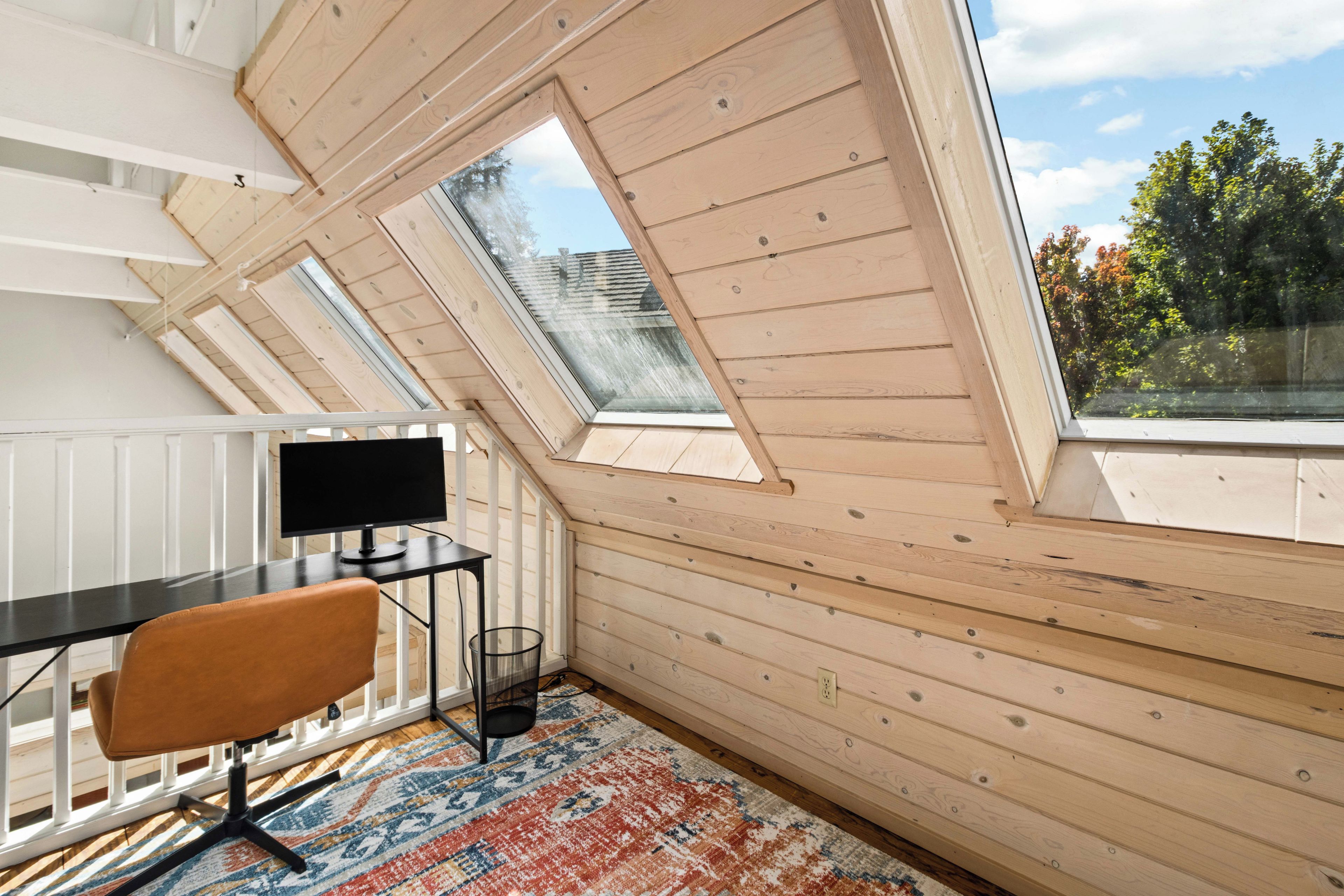 Une pièce avec un plafond en pente, un bureau, une chaise, un téléviseur et une fenêtre donnant sur les arbres.