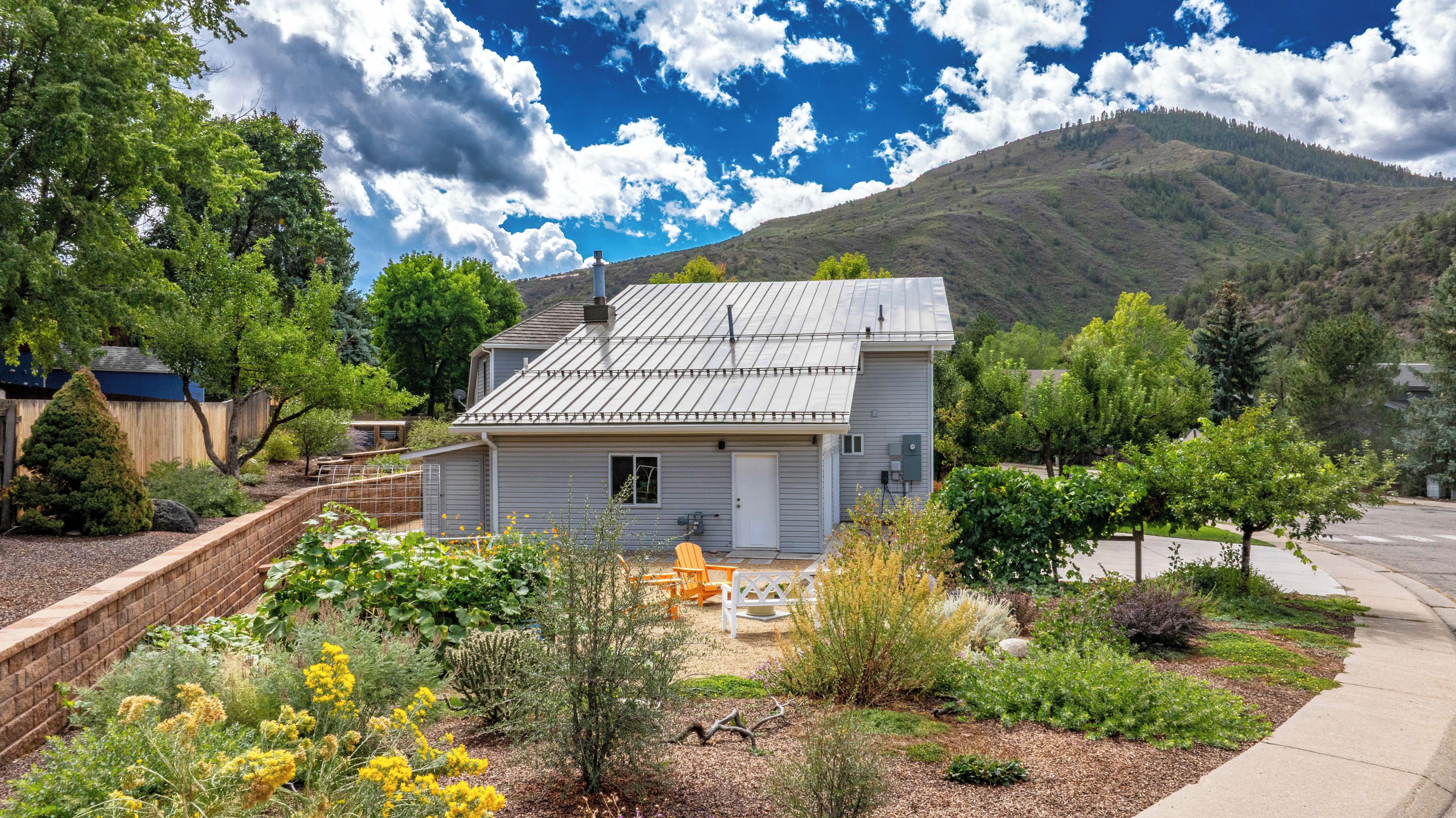 A single-story house with a metal roof, surrounded by a garden with various plants and a small patio area.