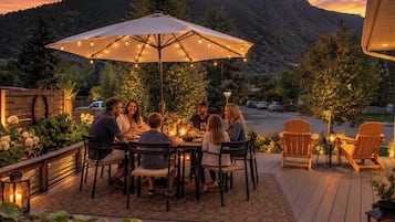 A group of people dining at an outdoor table with a large umbrella, surrounded by wooden deck and potted plants, with a mountainous backdrop.