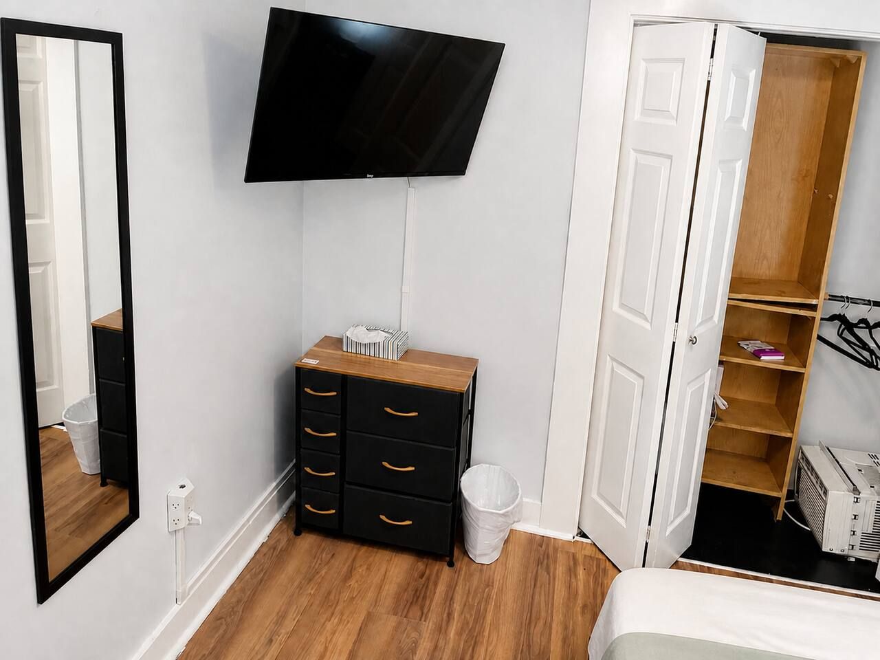Un dormitorio con una televisión de pantalla plana montada en la pared, un aparador de madera con cajones, un cubo de basura blanco y un armario con las puertas abiertas.