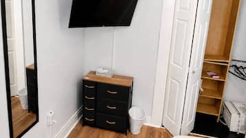 A bedroom with a flat-screen TV mounted on the wall, a wooden dresser with drawers, a white trash bin, and a wardrobe with open doors.