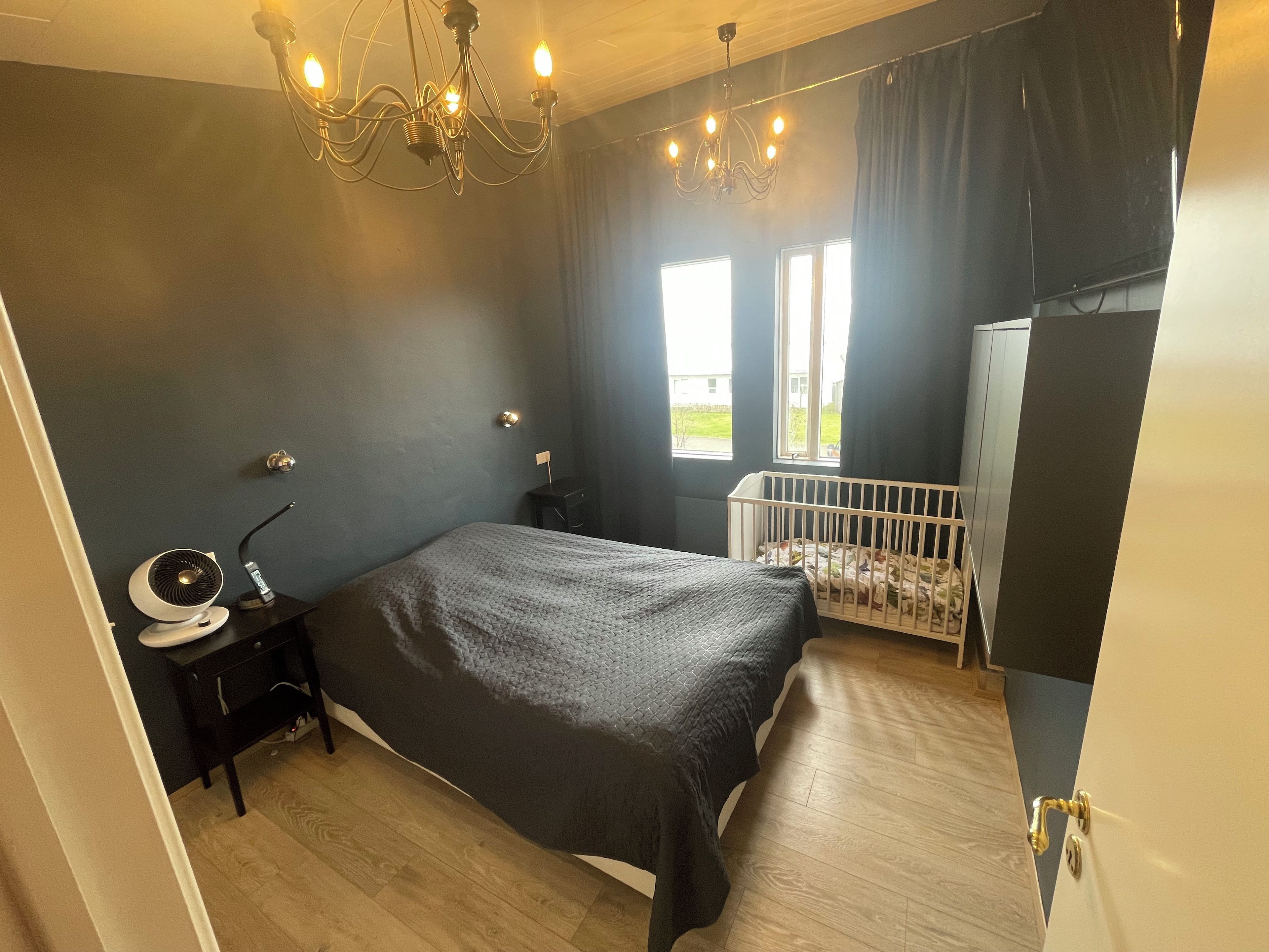 A bedroom with a bed, a crib, a bedside table with a lamp, a wall-mounted light fixture, and a window with curtains.