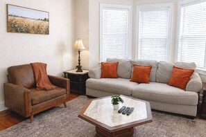Ein Wohnzimmer mit einem braunen Ledersessel, einem beigen Sofa mit orangenen Kissen, einem kleinen Tisch mit einer Lampe und einem Gemälde an der Wand.