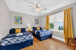 A hotel room with two beds, a ceiling fan, a window with yellow curtains, and artwork on the walls.