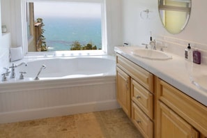 A bathroom with a bathtub, a sink, wooden cabinets, and a large window offering a view of the sea. - Serene Coastal Retreat Clark's Cove - Holiday Home (Jenner)