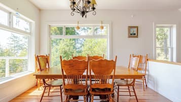 A dining room with a large table and six chairs, large windows with a view of trees, and a chandelier.