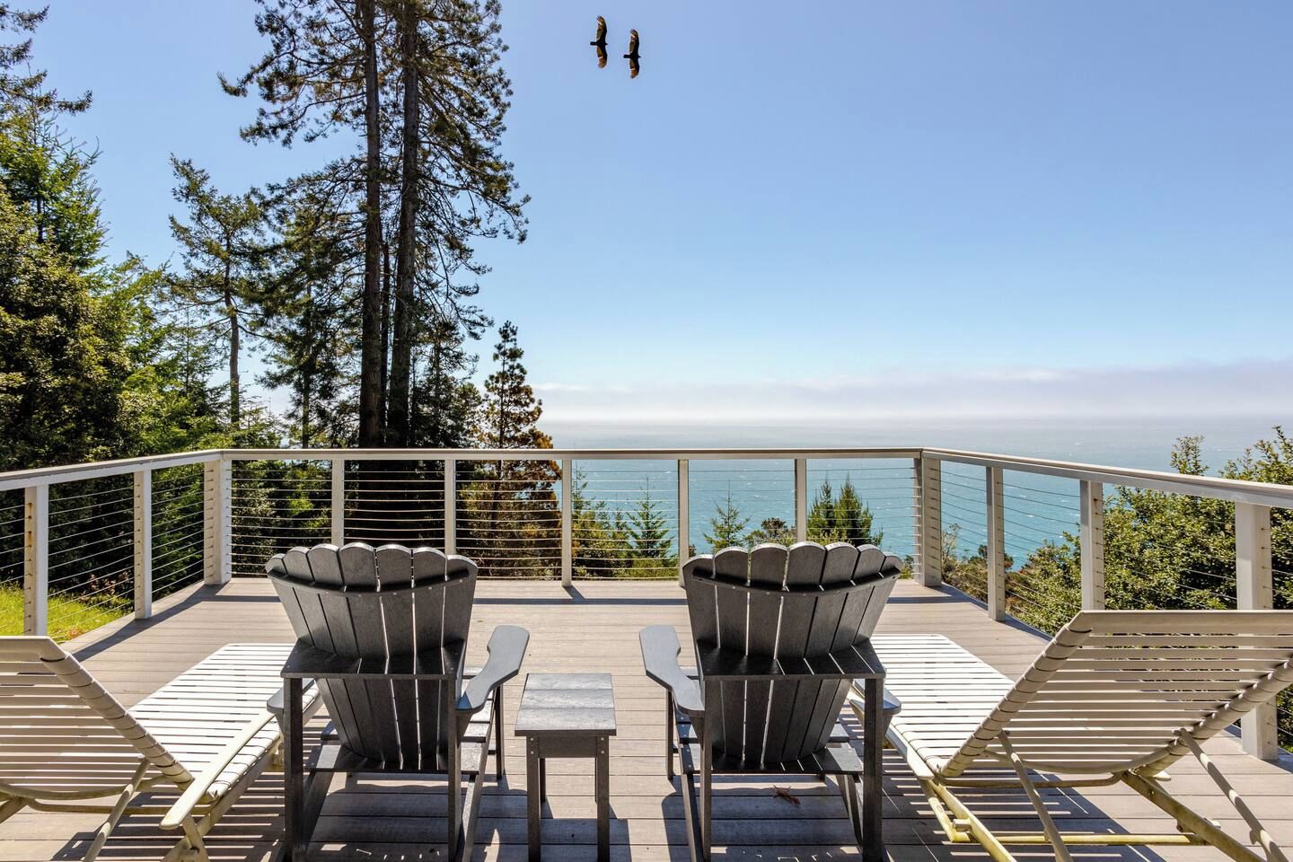 A deck with two Adirondack chairs, a small table, and a railing overlooking a body of water with trees in the background.