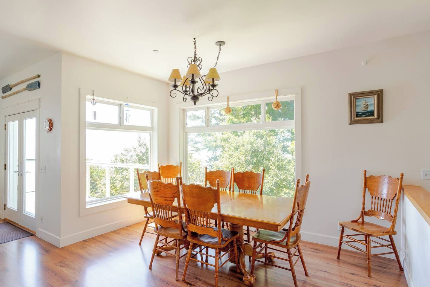 Comedor con una mesa grande y sillas, una araña y ventanas amplias con vistas a vegetación.