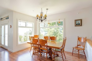 A dining room with a large table and chairs, a chandelier, and large windows with a view of greenery.