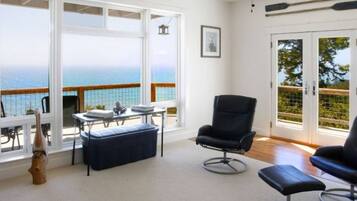A room with a view of the sea, a black chair, a table with a bench, and a framed picture on the wall.