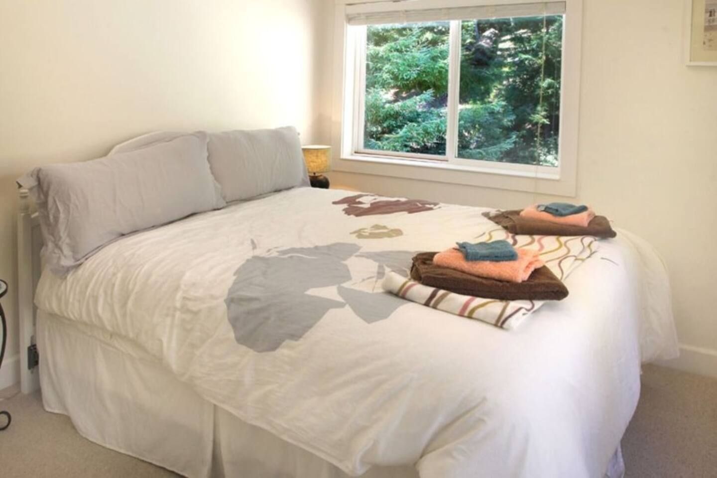 Een netjes opgemaakt bed met een witte deken en kussens, een raam met uitzicht op groen, en een bedlampje.
