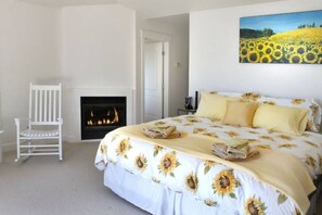 Un dormitorio con una cama, una silla blanca, una chimenea y un cuadro de girasoles.