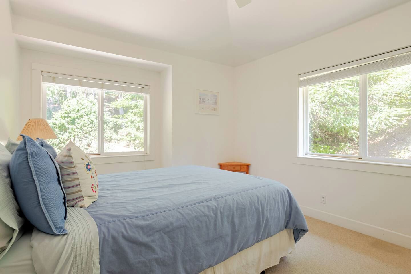 Un dormitorio con cama, dos ventanas, una mesita de noche e interruptor de luz en la pared.