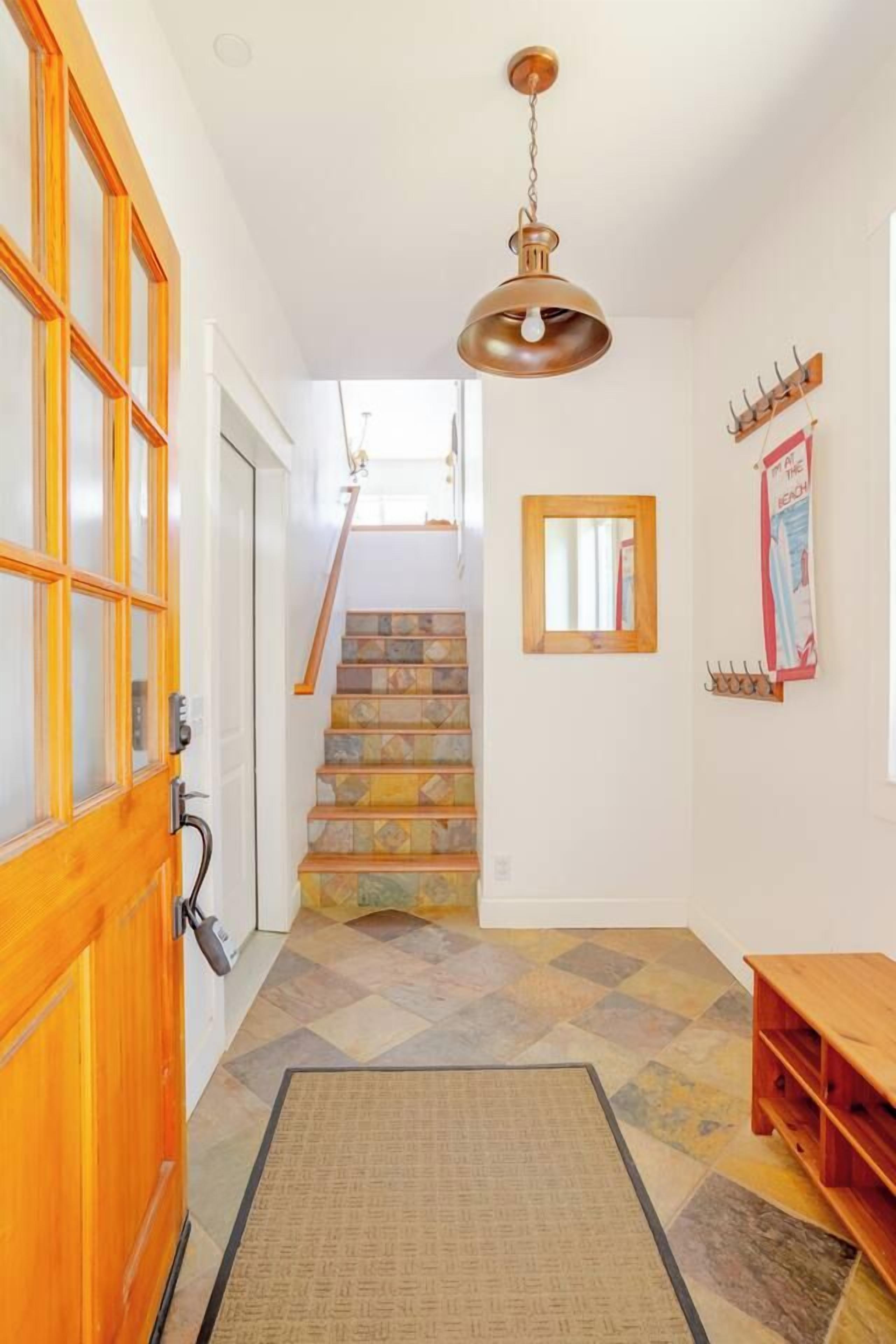 A hallway with a wooden door, a staircase with patterned tiles, a hanging pendant light, and a wall-mounted mirror.