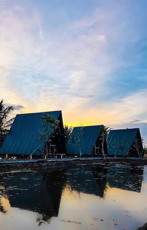 A row of buildings with sloped roofs reflecting on calm water, likely a lakeside or riverside property. - Ruhunumaya Nature Resort (Hambantota)