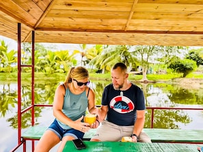 Two people sitting at a wooden table under a shelter with a view of a water body and greenery. - Ruhunumaya Nature Resort (Hambantota)
