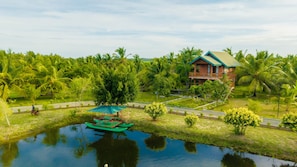 A house with a balcony, surrounded by lush greenery and a calm water body.