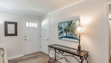 A hallway with a wooden floor, white walls, a glass-top console table with a lamp, and a framed abstract painting.