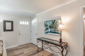 A hallway with a wooden floor, white walls, a glass-top console table with a lamp, and a framed abstract painting. - Johnson Oaks (Mobile)