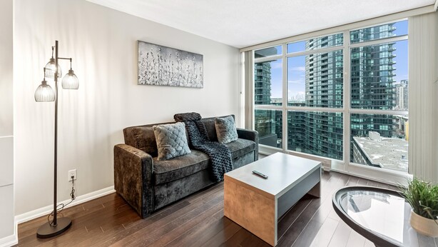 A modern living room with a sofa, a coffee table, a floor lamp, and a large window with a city view. - StayToronto - Fort York Prime Suites (Toronto)