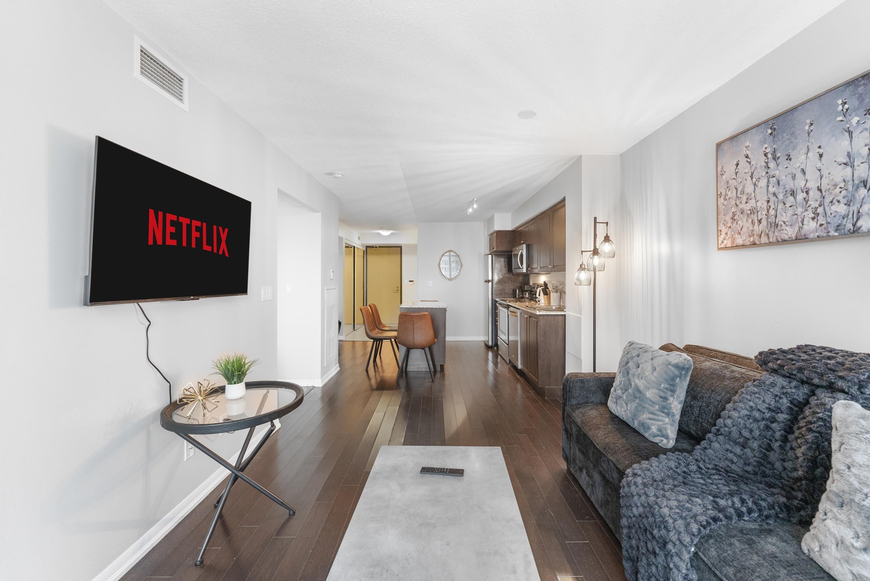 A modern living room with a flat-screen TV, a sofa, a coffee table, and a dining area in the background.