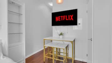 Una habitación con pared blanca, un televisor montado mostrando 'NETFLIX', una mesita pequeña con marco dorado y una puerta blanca.