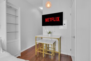 Una habitación con pared blanca, un televisor montado mostrando 'NETFLIX', una mesita pequeña con marco dorado y una puerta blanca.