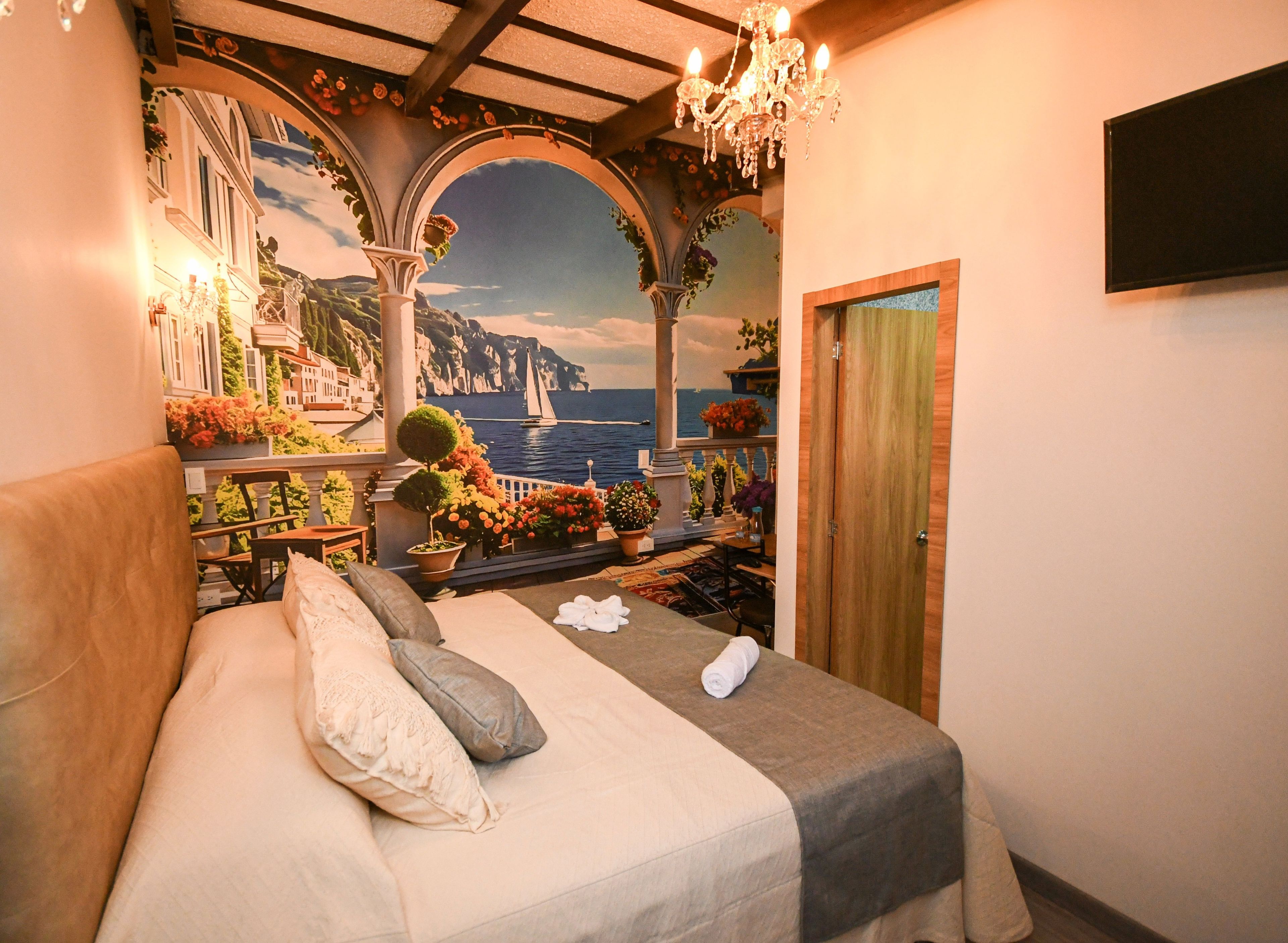 A bedroom with a bed, a wooden door, a chandelier, and a mural of a coastal scene.