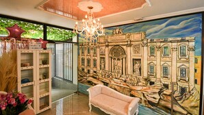 A lobby with a chandelier, a large mural of a building, a sofa, and a reception desk. - Hotel Fontana Di Trevi Boutique (Bogotá)