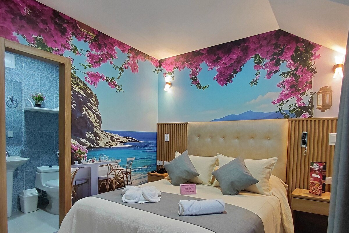 Un dormitorio con cama, mesitas de noche, baño con lavamanos, inodoro y ducha, y un mural con una escena de playa.