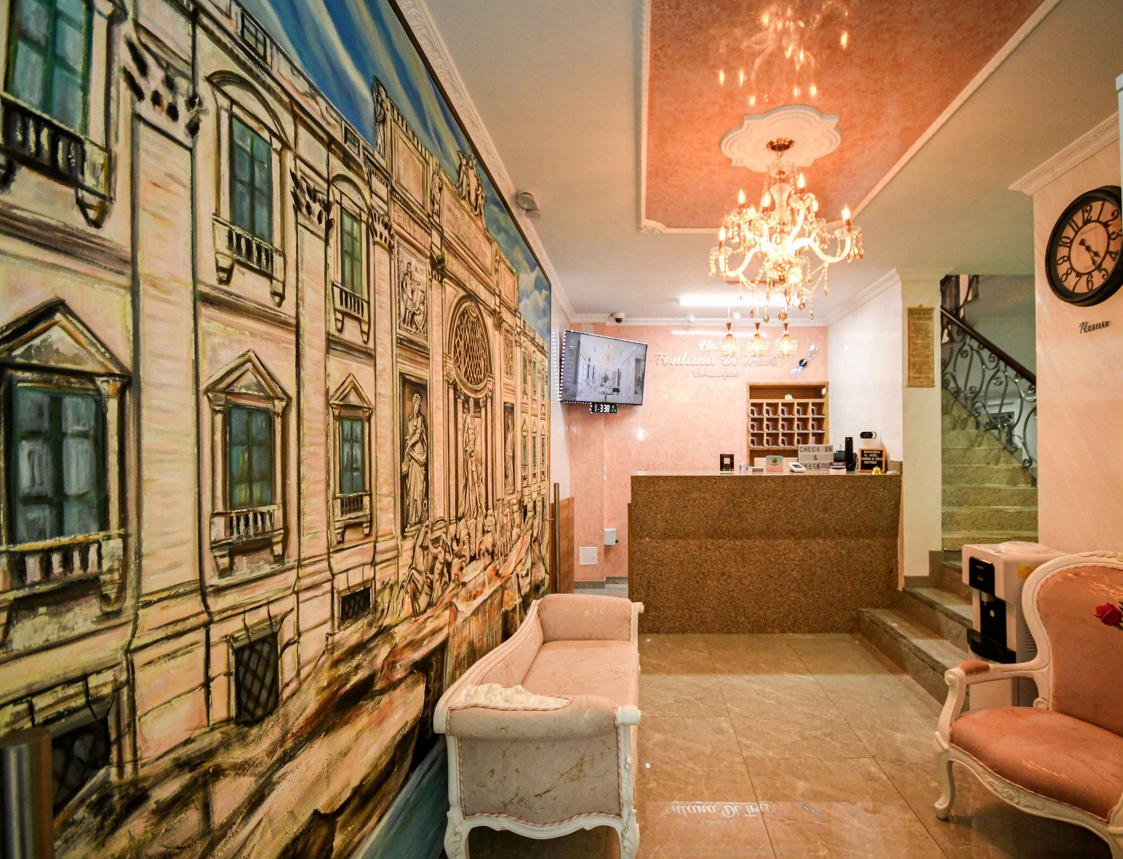 Een hotellobby met een grote muurschildering, een receptiebalie, een kroonluchter en een klok aan de muur.
