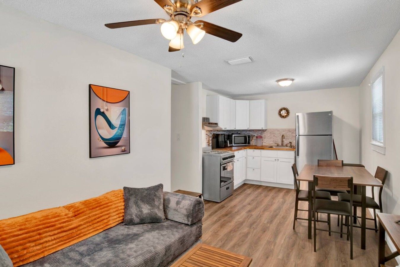 A modern living room with a sofa, a dining table with chairs, a kitchen area with appliances, and a ceiling fan.