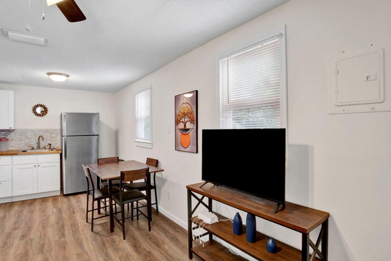 A compact kitchen with a refrigerator, a dining table with chairs, a flat-screen TV on a stand, and a window with blinds.