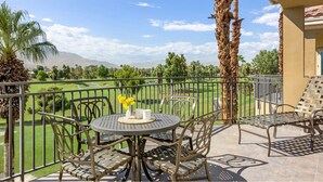 A balcony with a round table and chairs, a small vase with yellow flowers, and a view of a golf course and mountains. - Marriott Vacation Club- large 2 bd 2ba unit (Palm Desert)