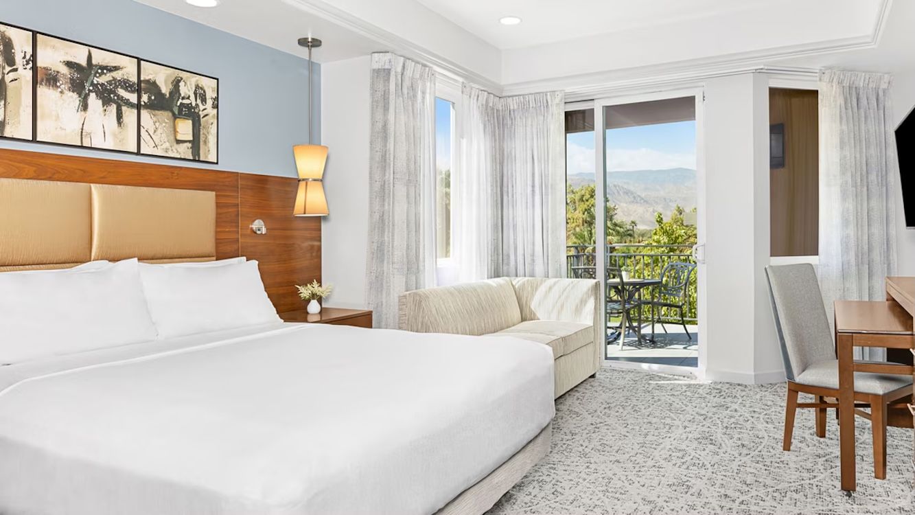 A hotel room with a large bed, a sofa, a dining table, and a view of the outdoors.