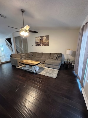 A living room with a sectional sofa, a coffee table, a ceiling fan, a painting, and a lamp.