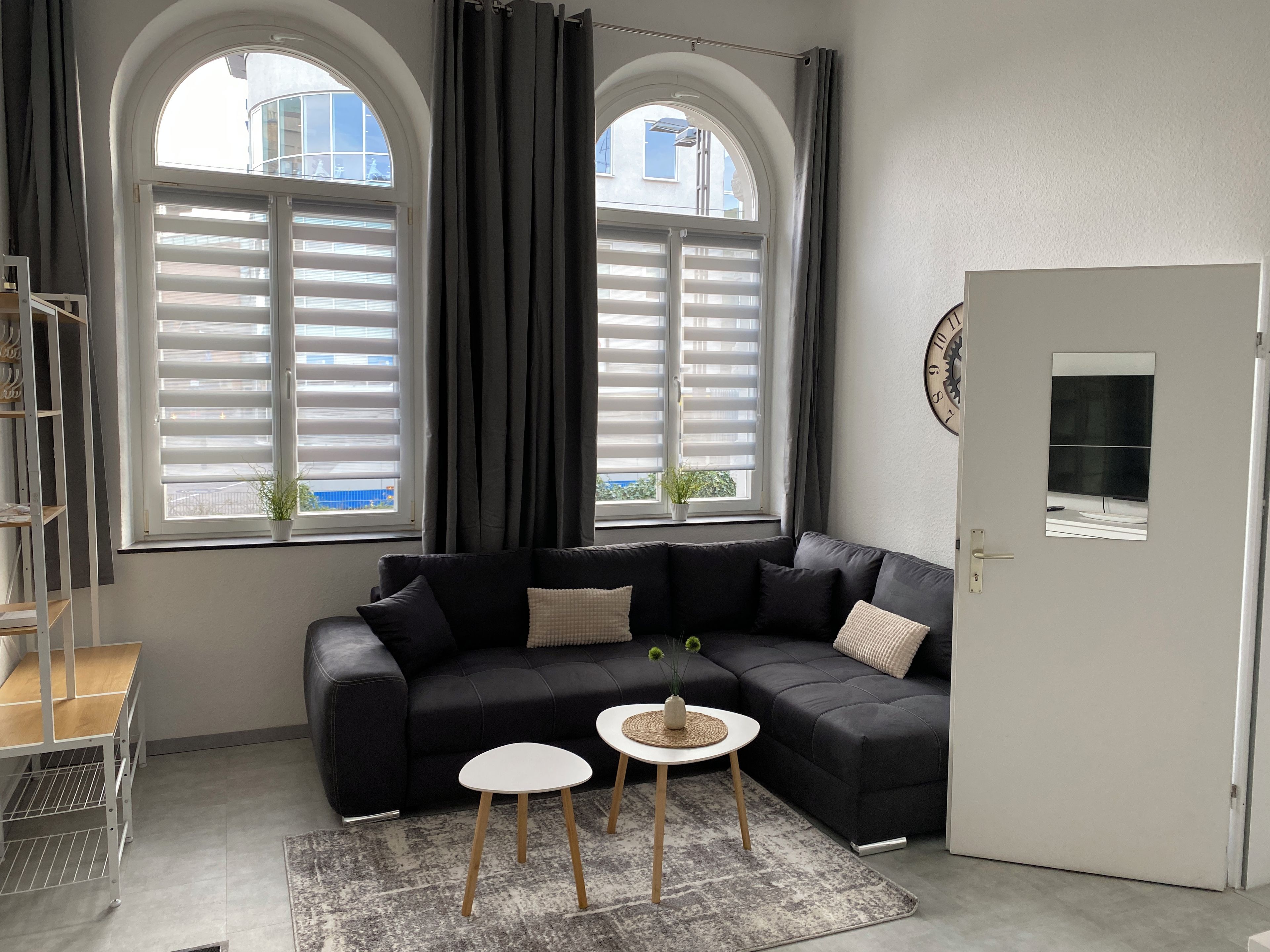 Ein modernes Wohnzimmer mit einem Ecksofa, einem kleinen runden Tisch, einer Topfpflanze und einer Tür mit einer Uhr.