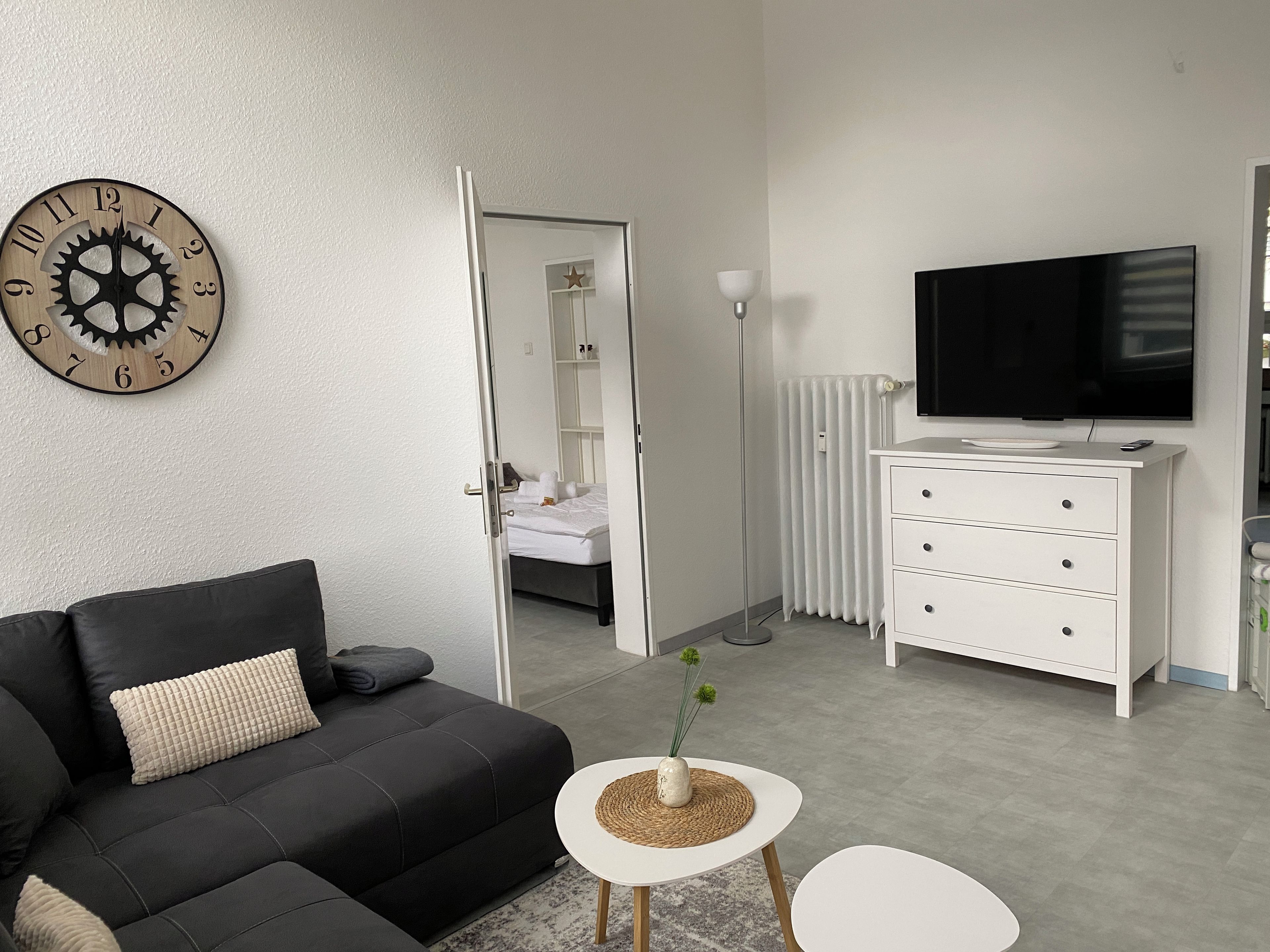 Ein modernes Hotelzimmer mit einer schwarzen Couch, einem weißen Schminktisch, einem Flachbildfernseher und einer Holz-Wanduhr.
