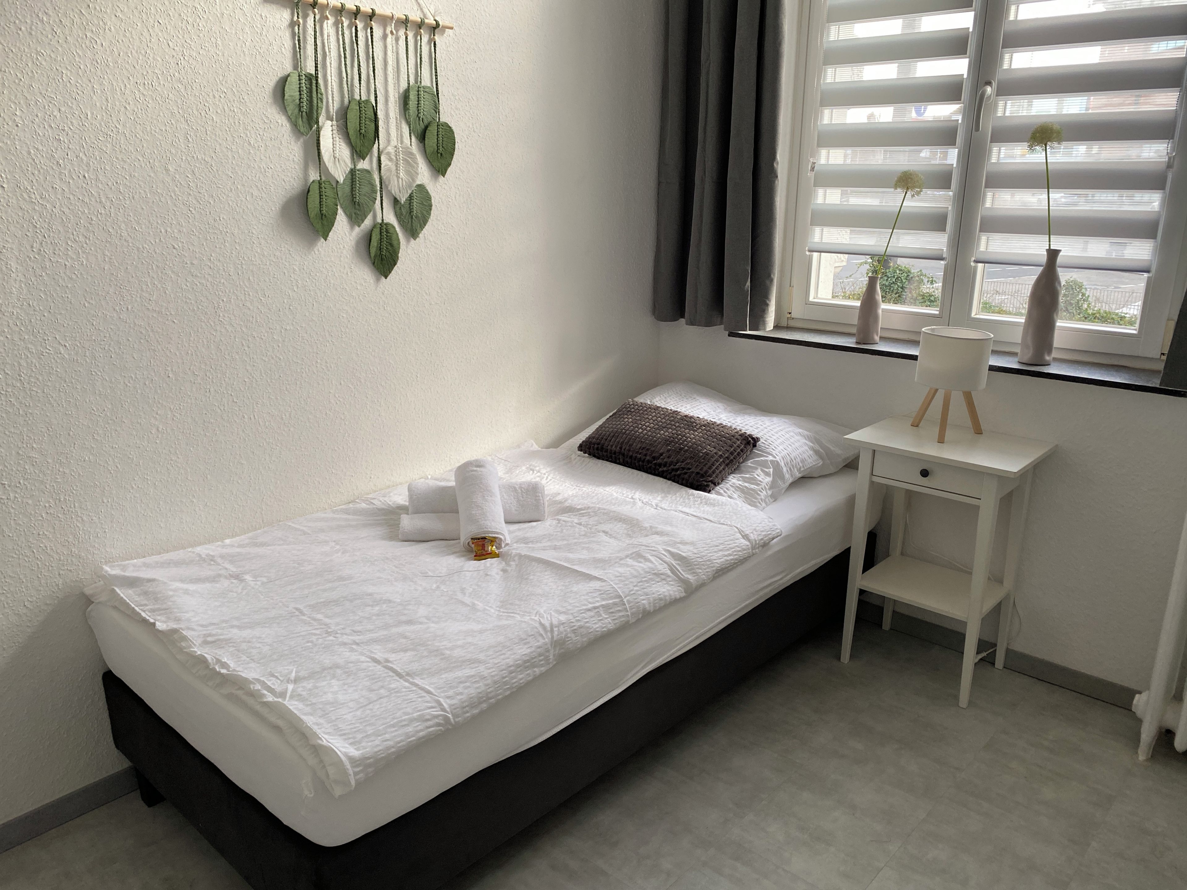 A single bed with white linens, a bedside table with a lamp, and a window with blinds.