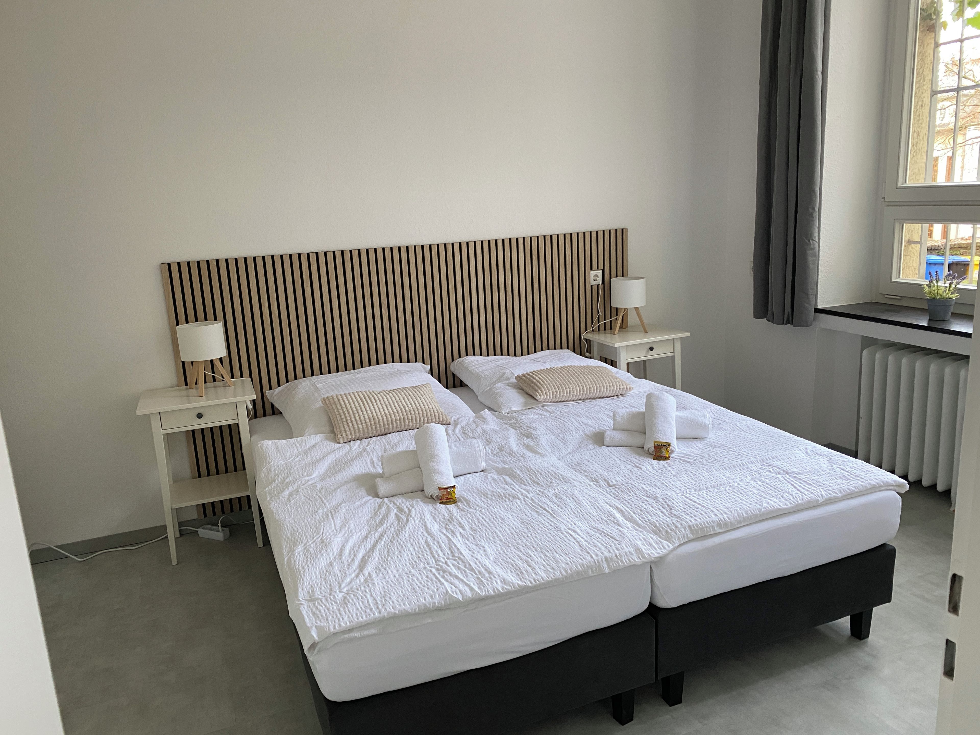 Ein Doppelbett mit weißer Bettwäsche, zwei Nachttische mit Lampen, ein gestreifter Kopfteil und ein Fenster mit Vorhängen.