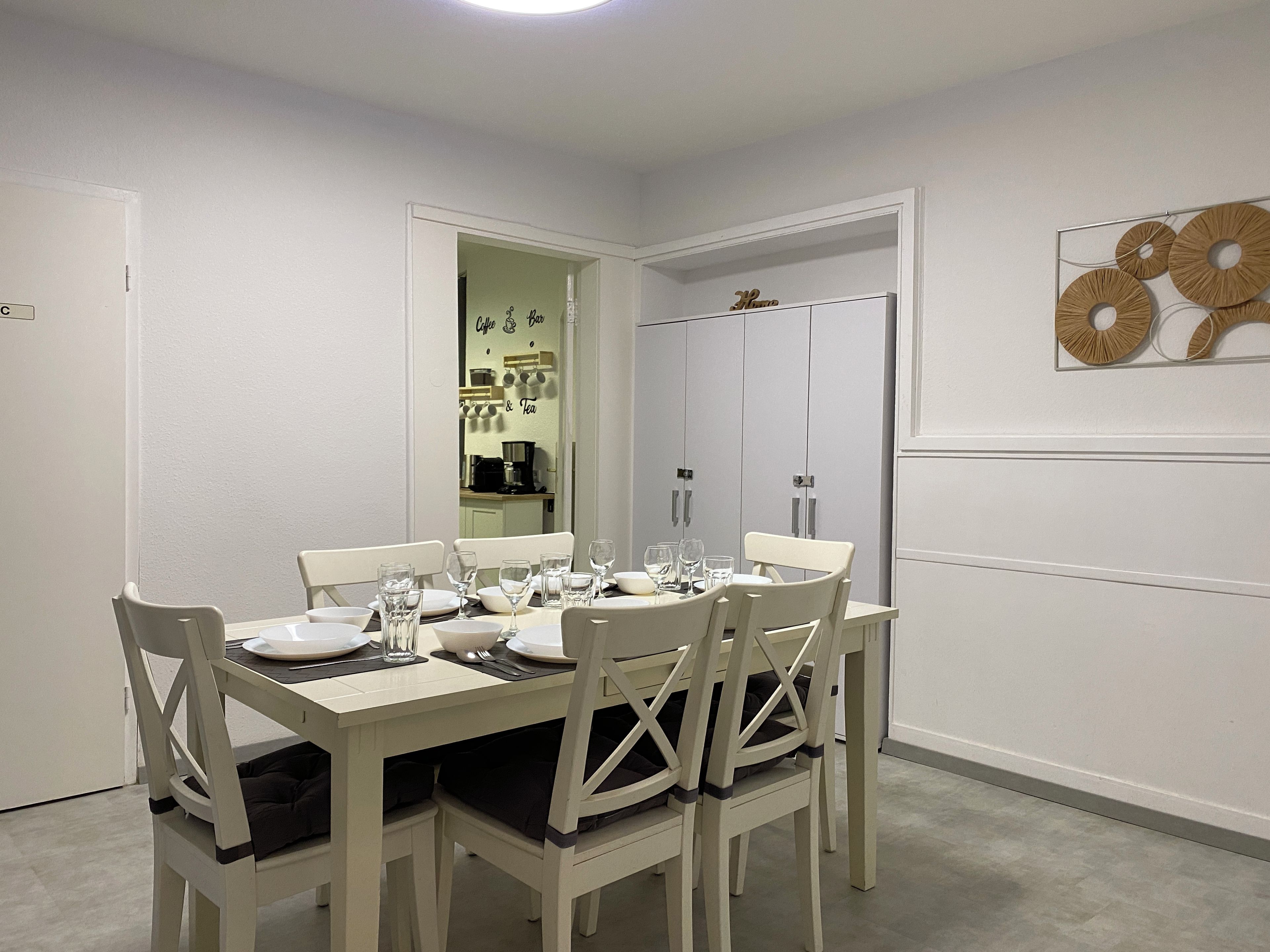 Ein Essbereich mit einem weißen Tisch, gedeckt für vier Personen, Stühle mit Kissen, einem Spiegel, der in eine Küchenzeile blickt, und einer Wand mit einem runden Dekorationsobjekt.