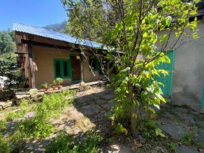 A rustic house with a corrugated metal roof, a wooden porch, and a small garden area with potted plants. - Tranquil 1-bedroom Mud House in charming Bashisht perfect for getaway (Bashisht)