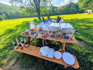 Disposição para refeições ao ar livre com uma mesa de madeira, garrafas e alimentos.
