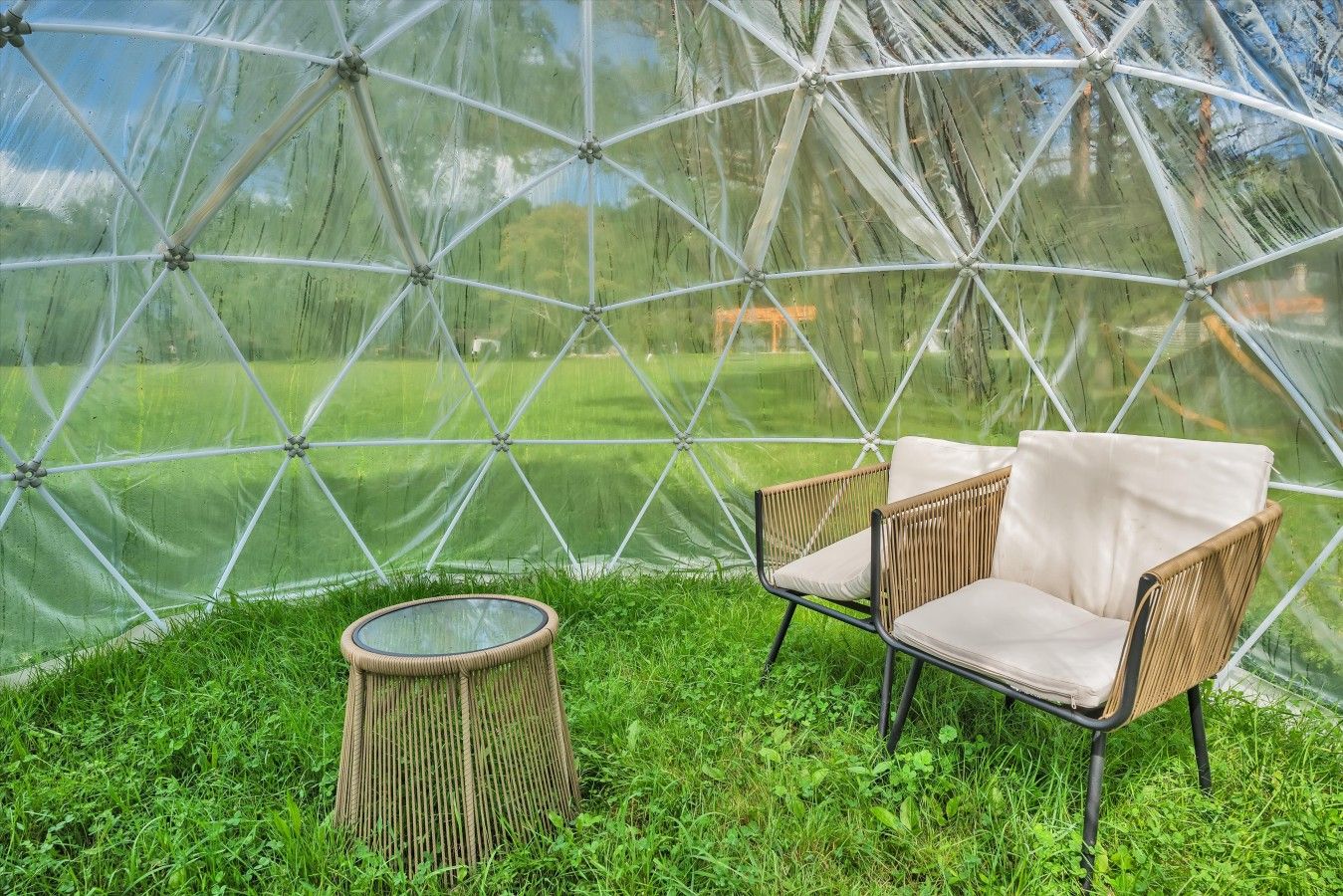 A transparent geodesic dome structure with a grassy interior, two wicker chairs, and a small round table.
