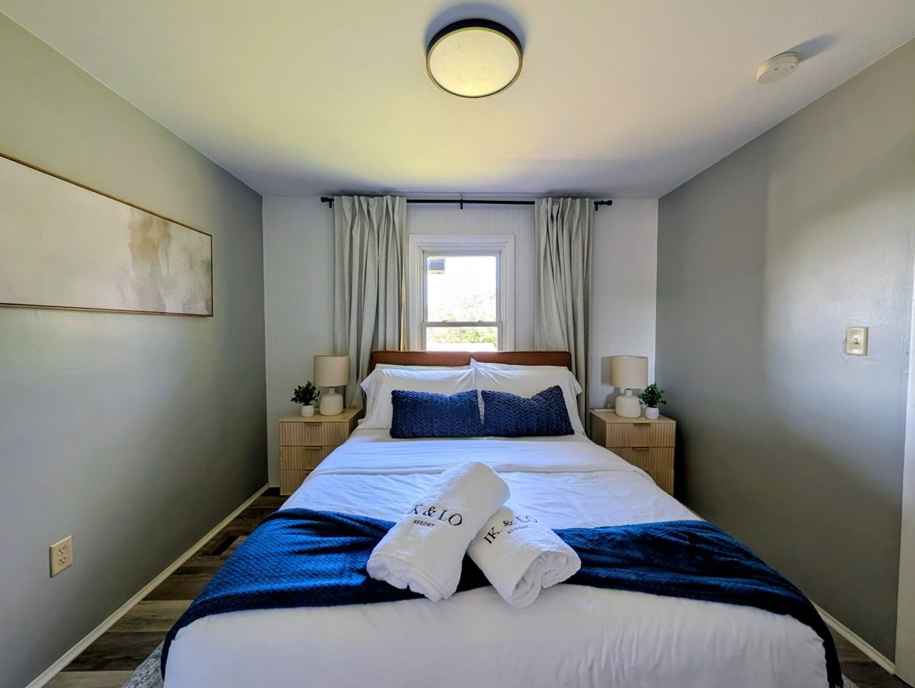 A hotel room with a bed, two bedside tables, a window with curtains, and a wall-mounted light fixture.