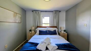 A hotel room with a bed, two bedside tables, a window with curtains, and a wall-mounted light fixture.