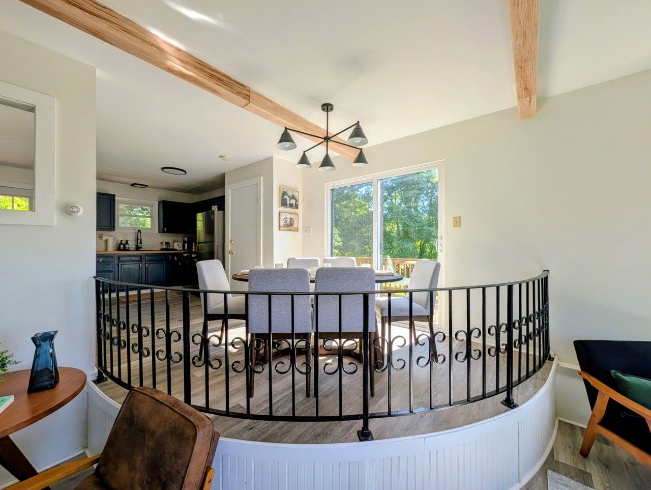 A dining area with a round table and chairs, a black metal railing, and a view of the kitchen and outdoors.