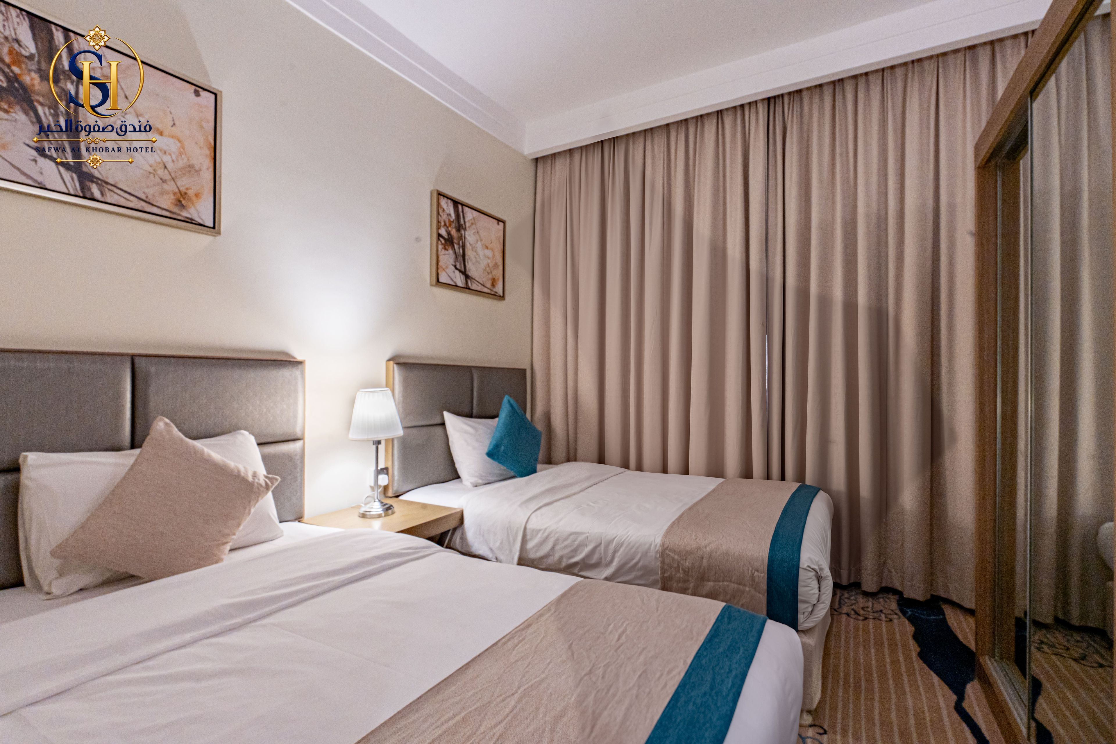 Una habitación de hotel con dos camas, una mesita de noche, una lámpara y un ventanal grande con cortinas.