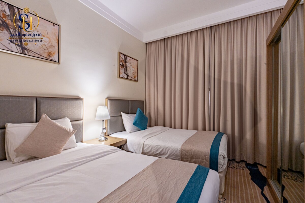 A hotel room with two beds, a nightstand, a lamp, and a large window with curtains.